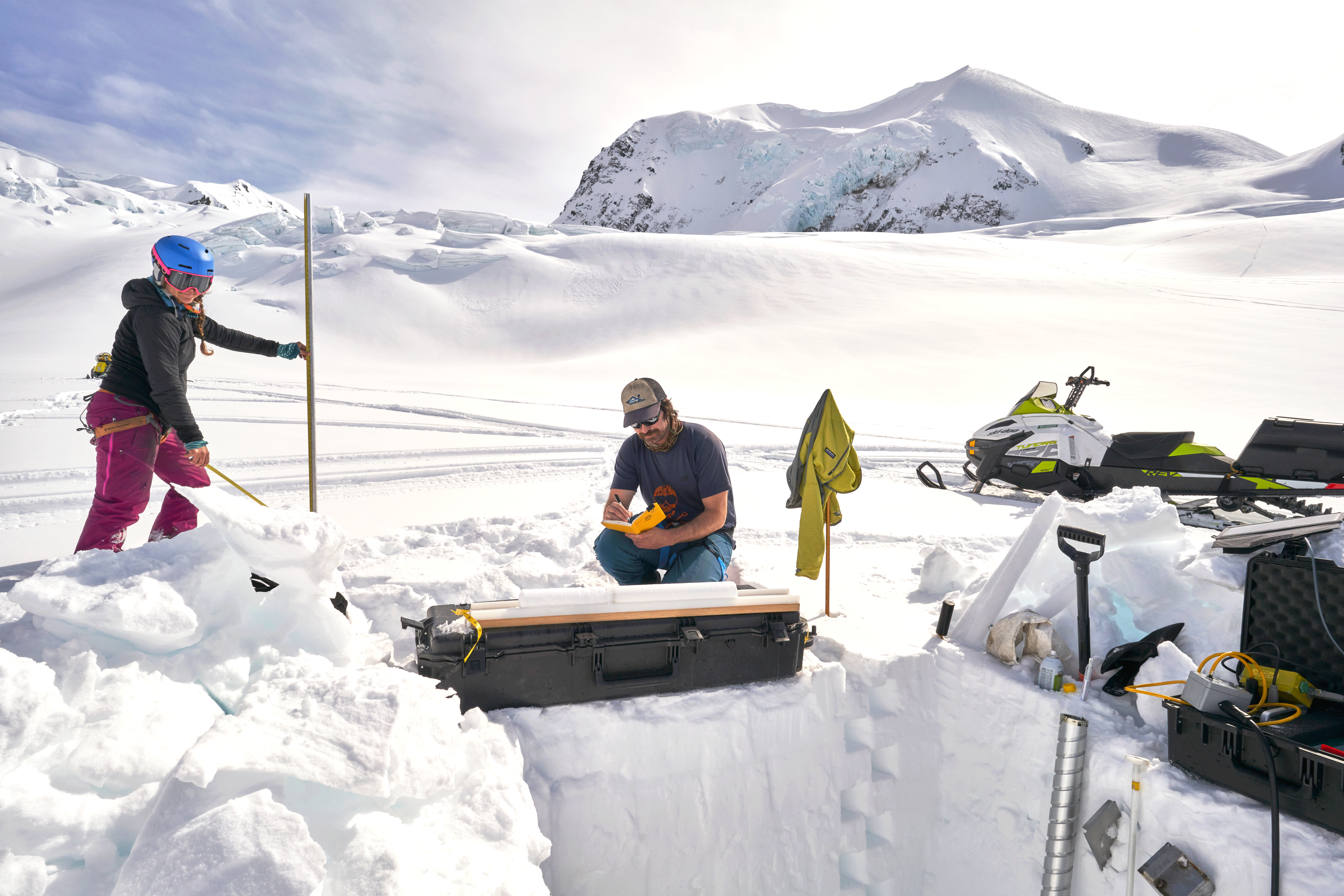 Scientists stand and kneel on a glacier behind a rectangular pit in the snow and several
ice cores, collecting snow accumulation and density data.